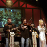 Dolly Parton in a white outfit singing on the Grand Ole Opry stage alongside fiddle, guitar and bass players