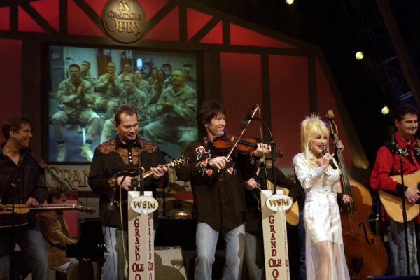 Dolly Parton in a white outfit singing on the Grand Ole Opry stage alongside fiddle, guitar and bass players